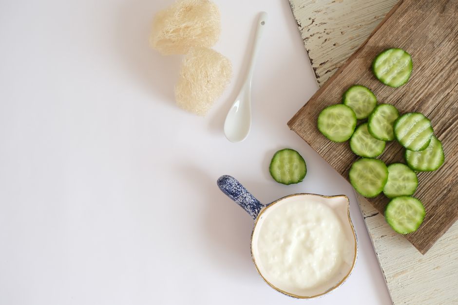 Flat lay of cucumber slices and creamy dairy in a ceramic jug on a wooden board and white background.