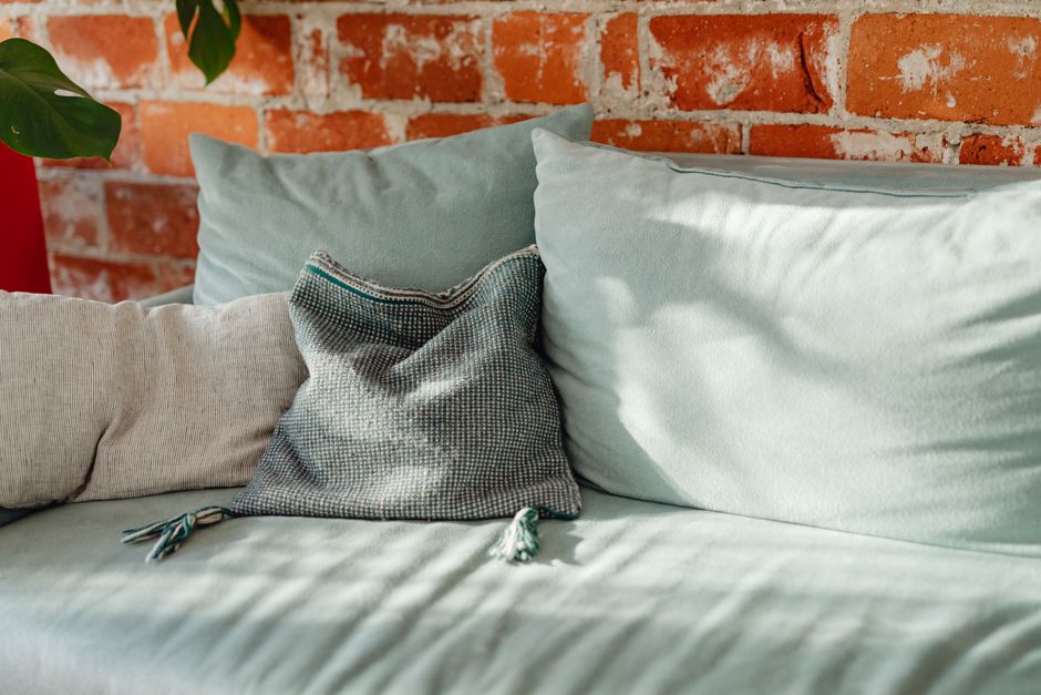 A stylish teal sofa with cushions against a rustic brick wall, perfect for cozy interiors.