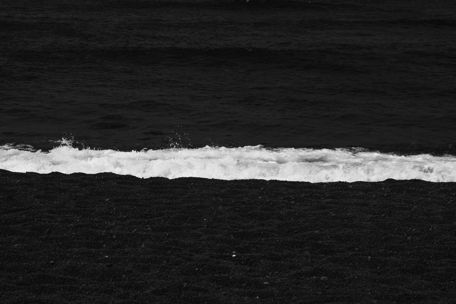 A serene black and white coastal photograph featuring a wave breaking on a pebble beach.