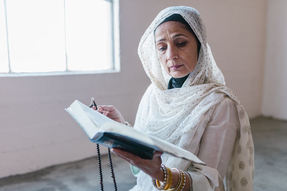 An elderly Muslim woman reads the Quran while holding prayer beads in a serene indoor setting.