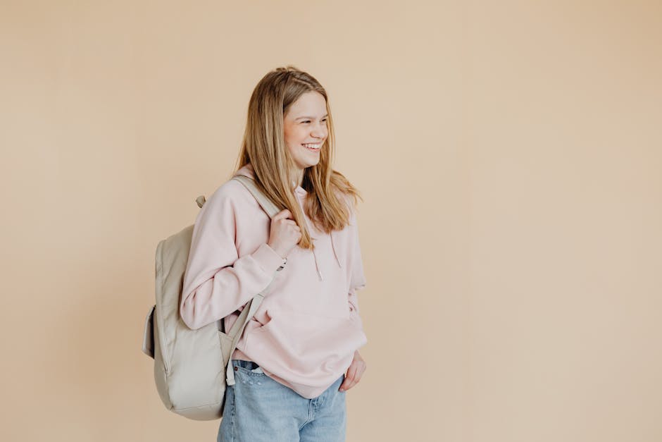Teenage girl smiling while wearing a hoodie and backpack against a beige background.
