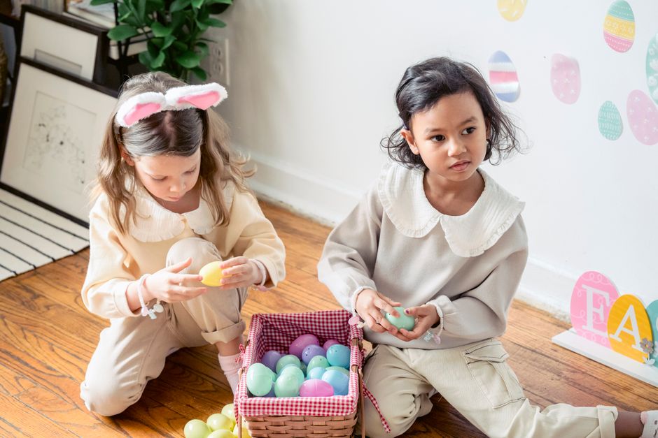 High angle of cute little multiethnic children in similar outfits sitting on wooden floor and playing with multicolored egg toys in wicker basket during Easter holiday