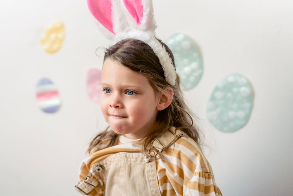 Adorable preschool girl with bunny ears headband standing near decorated wall with colorful eggs during Easter holiday celebration in room