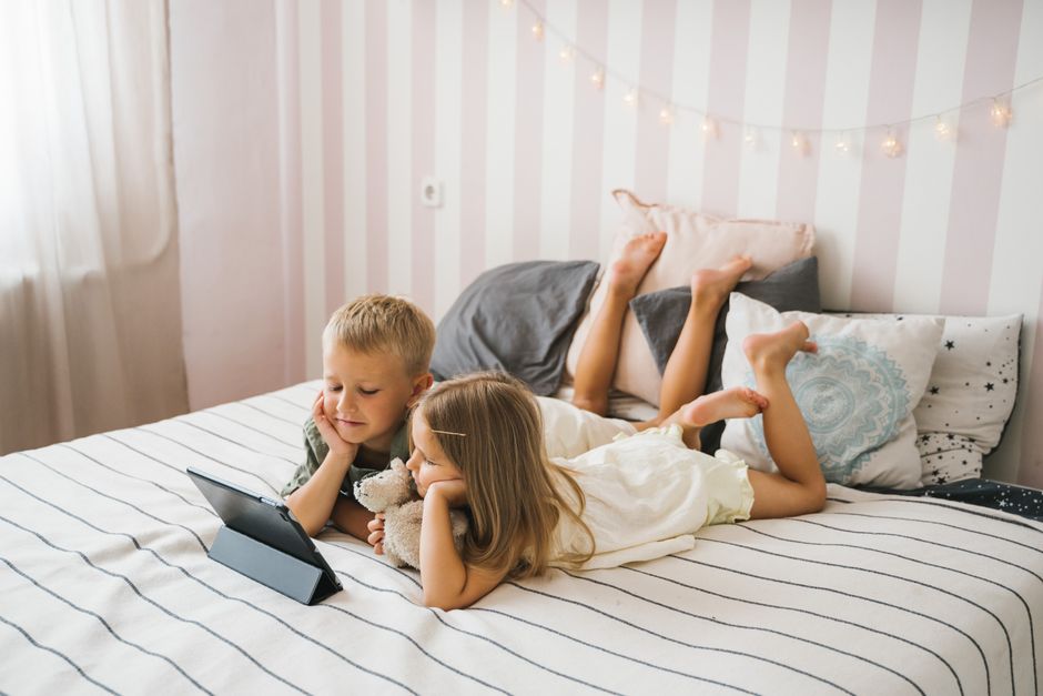 Young siblings lying on a cozy bed, engrossed in a tablet while enjoying a relaxed indoor setting.