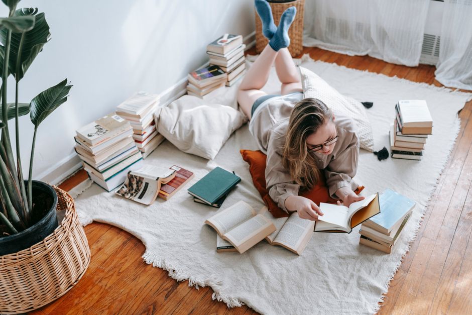 From above full body of focused young female reading book while lying on white textile with pile of literature in light room with flowerpots