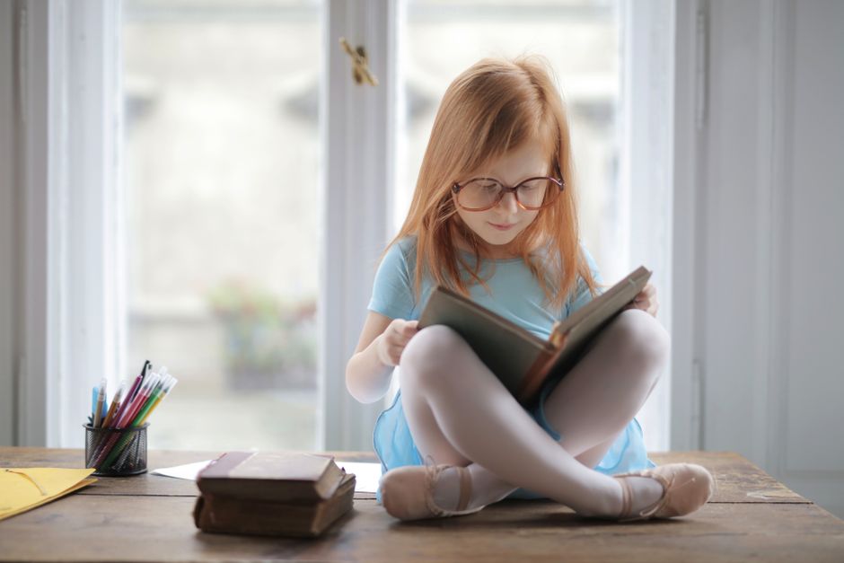 A young girl in glasses reads a book by a window, embodying knowledge and leisure in a cozy indoor setting.