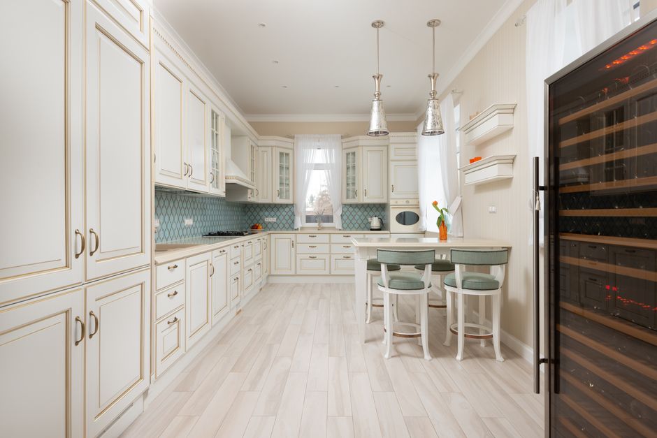 Interior of spacious modern kitchen with white cupboards and wooden cabinets near table with chairs under lamps next to shelves
