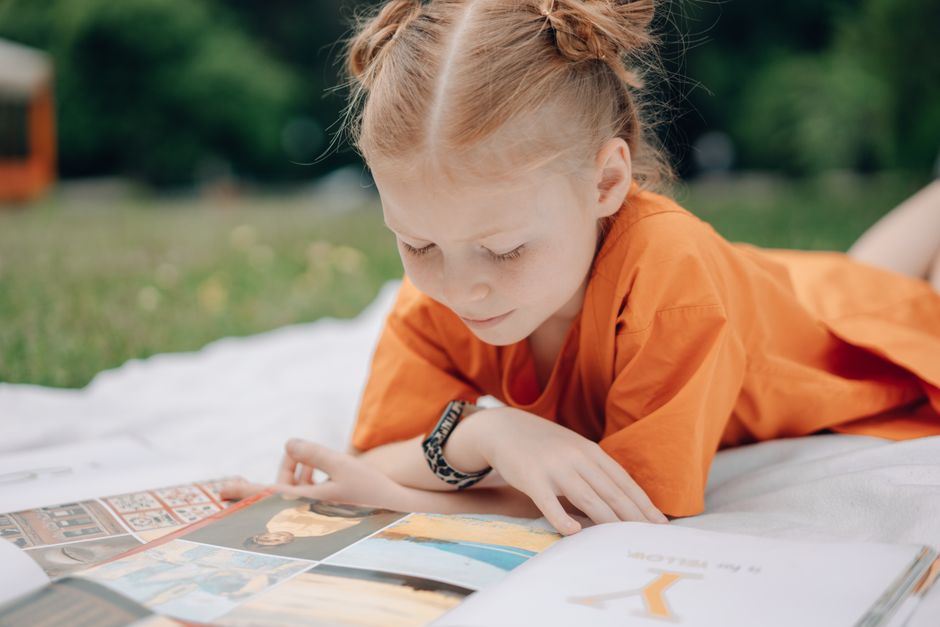 A young girl in an orange dress reads a book outdoors on a sunny day, enjoying a relaxed moment.