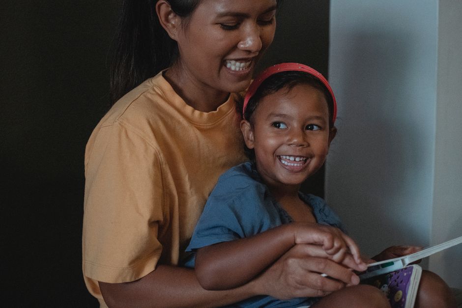 Smiling ethnic crop woman hugging playful girl while reading interesting story together near black wall during free time in apartment
