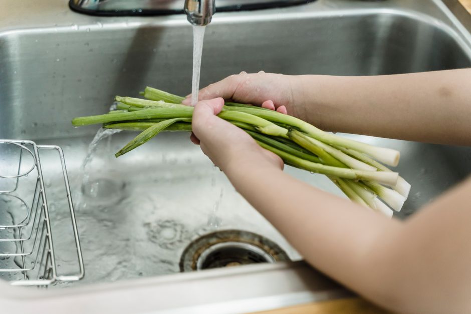 Hands washing fresh leeks under running water in a kitchen sink, ensuring hygiene and cleanliness.