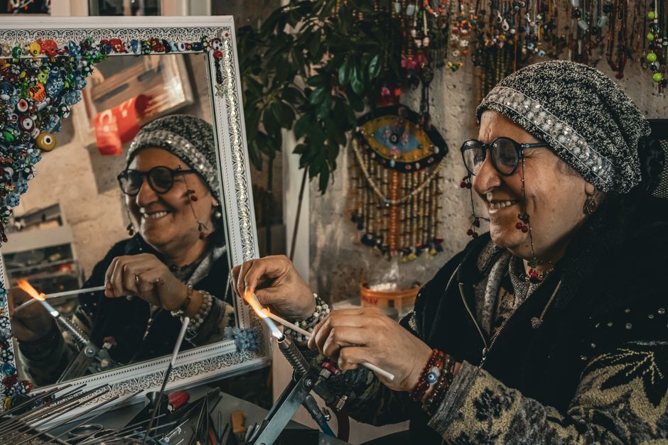 An artisan joyfully crafting glass jewelry, reflected in a decorative mirror, showcasing intricate bead designs.