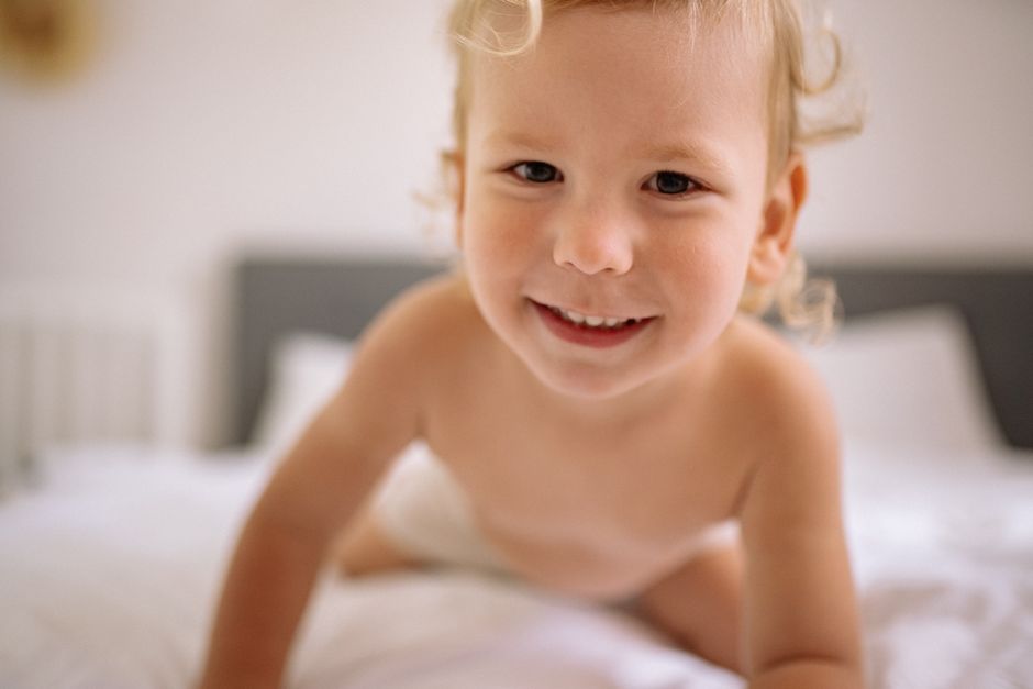 A cheerful toddler with blonde hair and brown eyes smiles while crawling on a bed indoors.