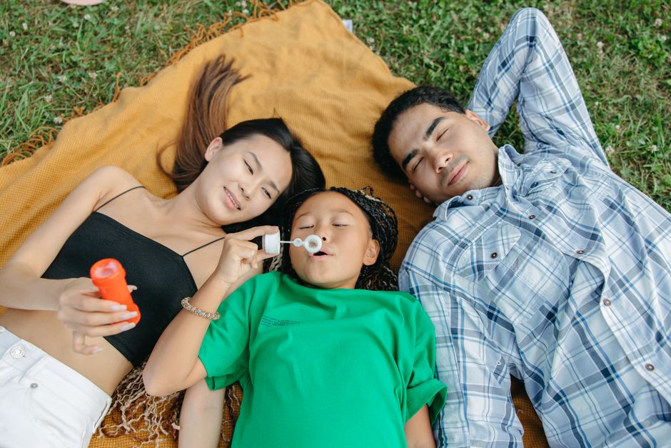 A family lying on a blanket outdoors enjoying a relaxing moment while a child blows bubbles.