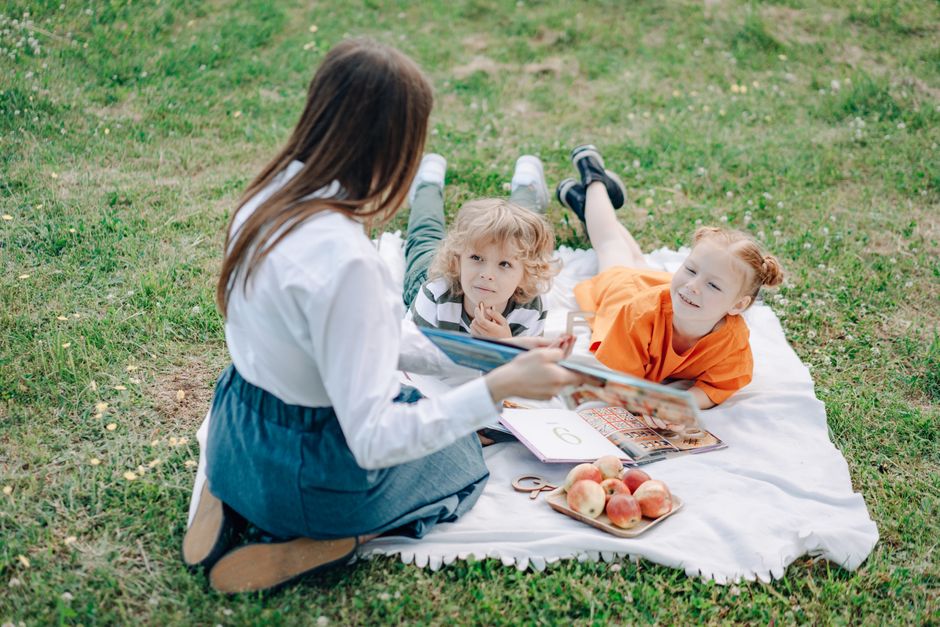 Children and teacher studying outdoors on a picnic mat with books and snacks.
