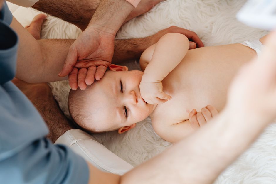 Cute baby lying on a soft surface, surrounded by caring hands, thumb sucking.