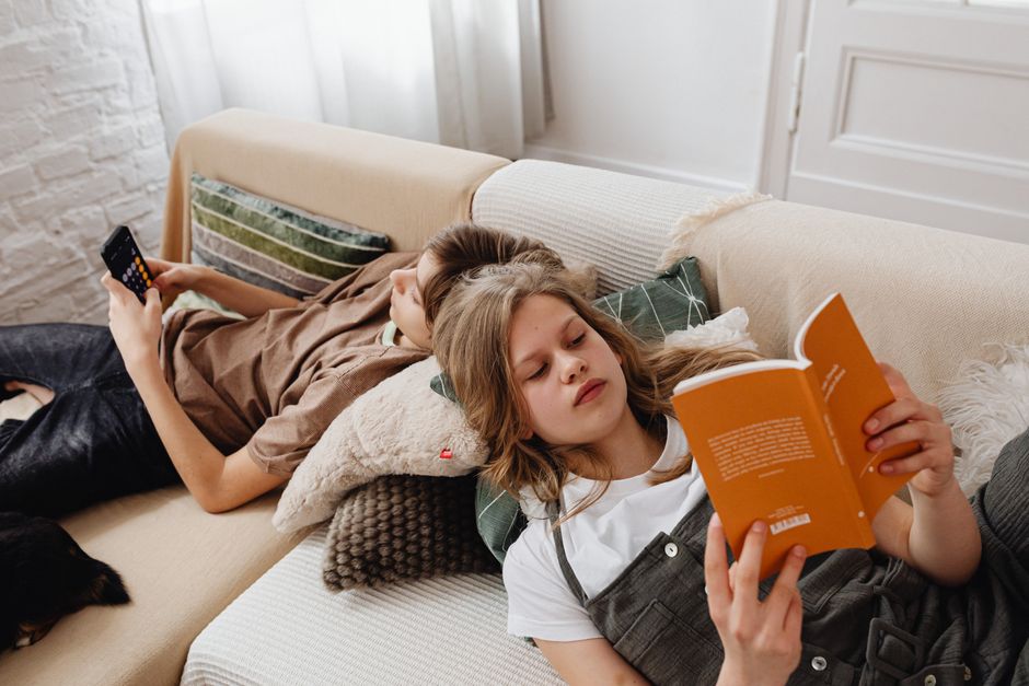 Siblings enjoying leisure time reading and using a phone on a cozy living room couch.