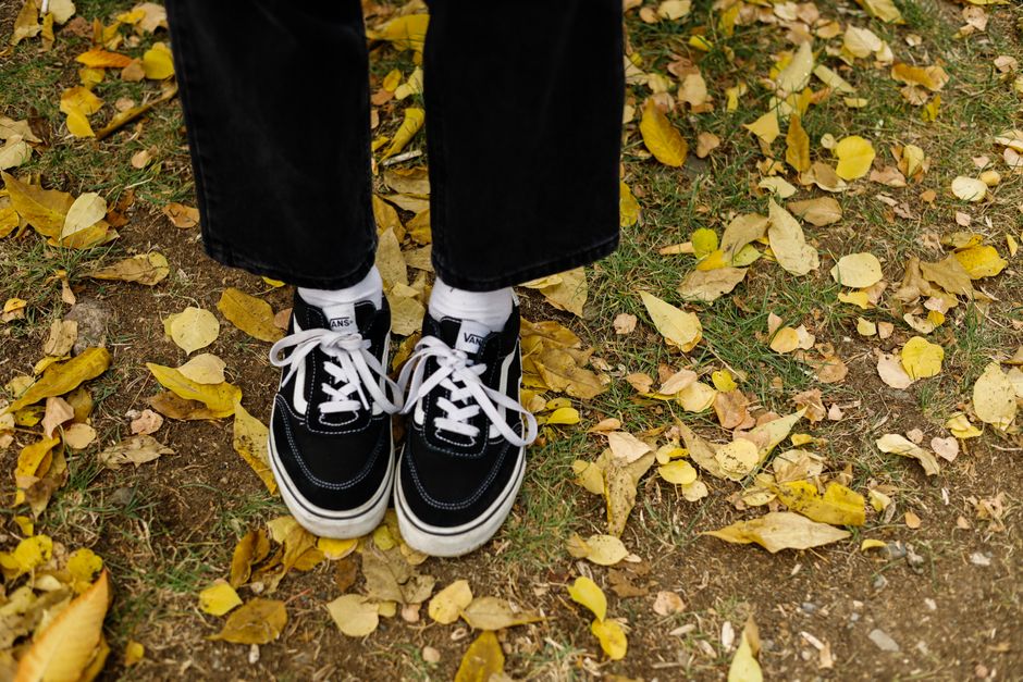 Low-angle shot of black sneakers with white laces on a grassy, leaf-covered path in autumn.