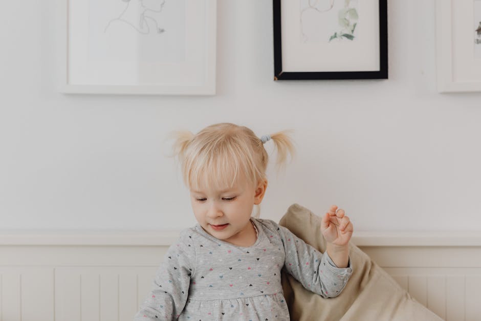 Cute blonde toddler in a gray dress plays indoors with framed art on the wall.