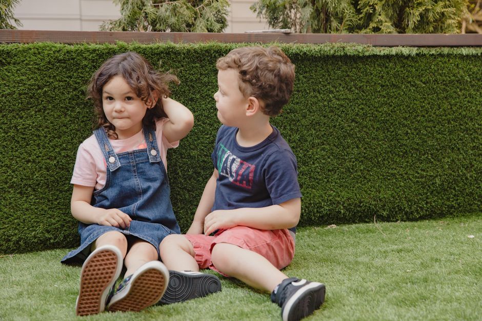 Two kids sitting on green grass outdoors, enjoying a playful moment during the day.