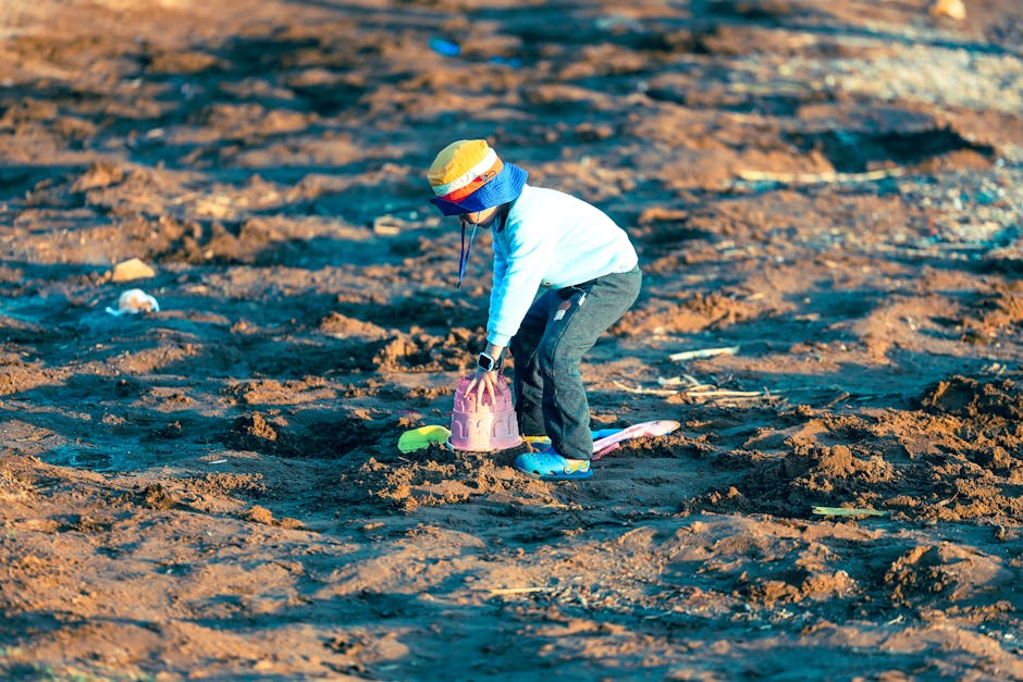 A young child wearing a colorful hat and blue shoes plays with sand toys outdoors on a sunny day.