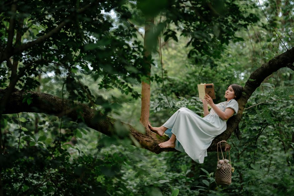 Young woman in a white dress reading on a tree branch amidst lush greenery, enjoying solitude.